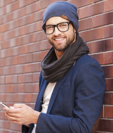 Man in beanie and scarf holding cell phone while standing in front of brick wall